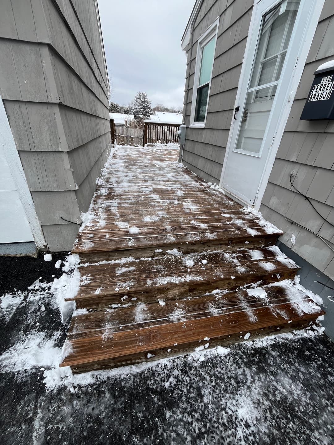 Snow-covered wooden steps leading to a house, with a cloudy sky in the background.